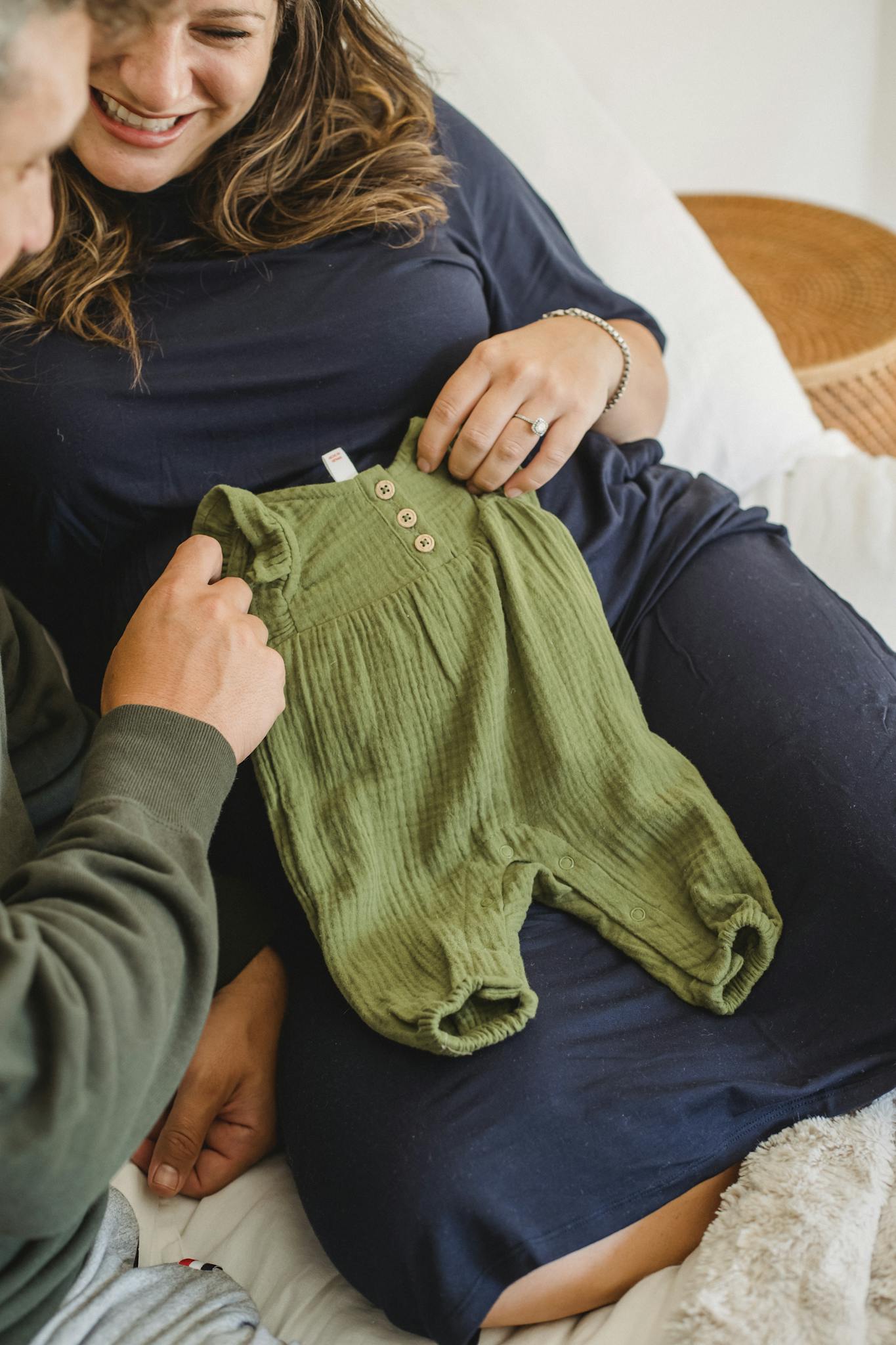 From above of crop unrecognizable man with smiling pregnant wife relaxing on bed and showing cute newborn baby clothes